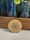 Cork coaster with a geometric design on a white surface next to decorative bottles and flowers.